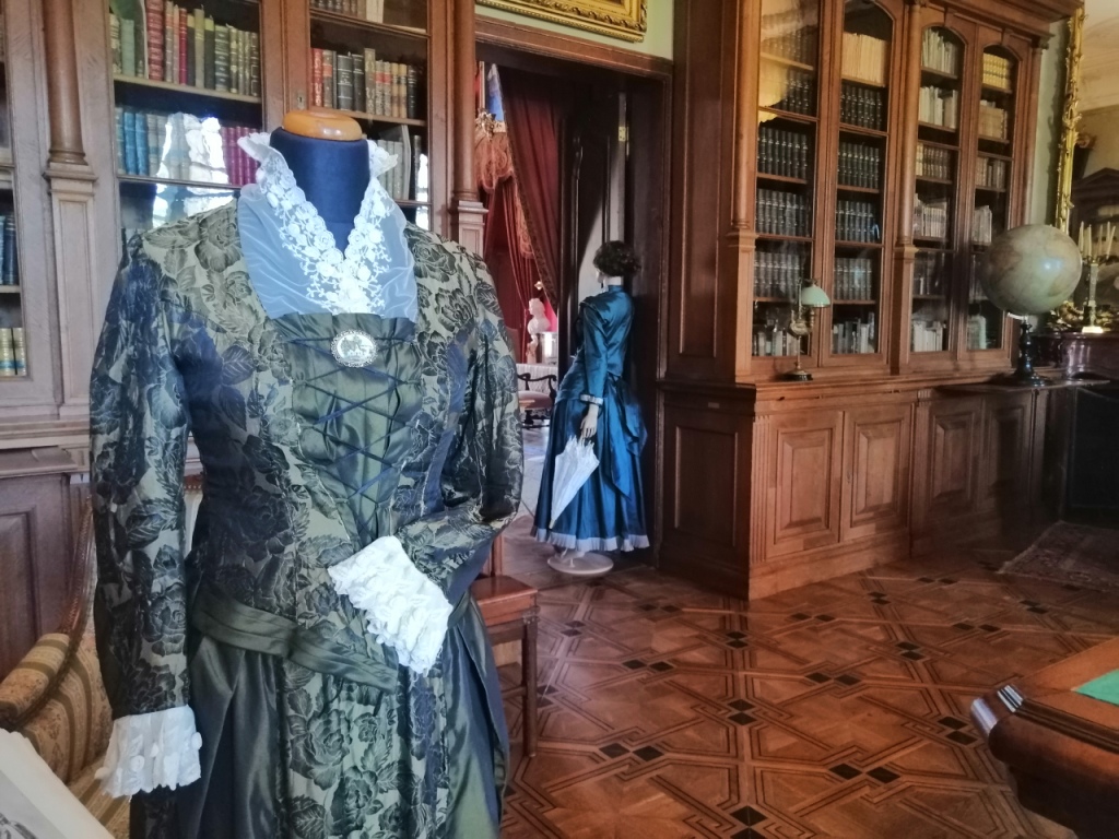 Wystawa "Modna w każdym wieku" Fotografia. Na tle drewnianych szaf wypełnionych książkami, dwie stylizowane dziewiętnastowieczne kreacje damskie zaprezentowane na manekinach. Na pierwszym planie suknia uszyta z oliwkowej tkaniny z wzorem czarnych delikatnych róż. Stanik sznurowany na piersiach, dekolt wypełniony szmizetką z delikatnego materiału wykończonego kwiatowym haftem. Identycznie wykończone są rękawy. Na drugim planie ukazana od tyłu suknia uszyta z materiału w kolorze morskim. Mankiety długich rękawów i dół sukni wykończone suknem w kolorze stalowym. W lewej dłoni manekina koronkowa parasolka.