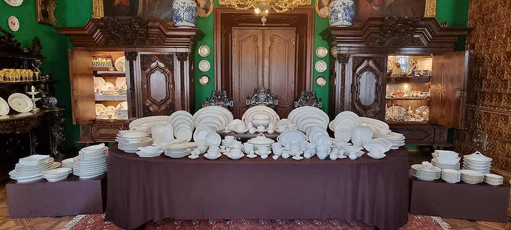 Jadalnia, ekspozycja do wystawy „Kredensie, otwórz się” Fotografia przedstawia pałacowe wnętrze. W centrum nakryty brązową tkaniną stół. Na nim porcelanowe naczynia w kolorze biały: półmiski, talerze, waza, filiżanki, kieliszki na jajka, maselniczki, solniczki, sosjerki. Po obu stronach stołu brązowe kubiki. Na nich białe porcelanowe naczynia ułożone w stosy. Po lewej stronie stołu fragment brązowego bufetu. Na nim talerze, elementy serwisu szklanego. Po prawej stronie stołu fragment pieca kaflowego w kolorze brązowym. Za stołem trzy brązowe krzesła, brązowe drzwi i dwie szafy w kolorze brązowym, otwarte w połowie. W szafie z lewej strony naczynia porcelanowe. W szafie z prawej strony naczynia srebrne.