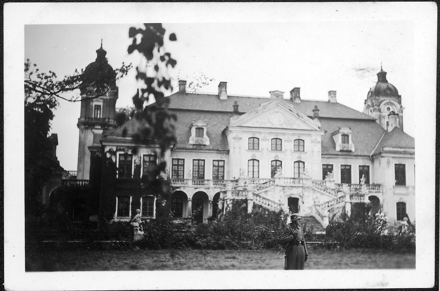 2. Pałac w Kozłówce, około 1940 roku; fotografia archiwalna; zbiory Muzeum Zamoyskich w Kozłówce, MPK/I/444 2. Fotografia archiwalna z lat 40. XX wieku przedstawia pałac w Kozłówce od strony ogrodu. Pałac jednopiętrowy, z wysuniętą częścią środkową , przy której duże schody oraz dwiema wieżami po obu stronach. Na tle pałacu stojący mężczyzna w mundurze żołnierza Wermachtu. Fotografia czarno-biała.