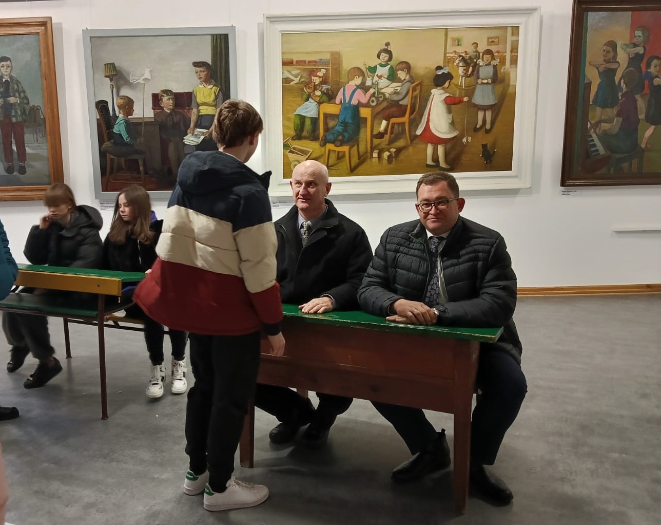 Mikołajki w Muzeum Zamoyskich w Kozłówce Na kolorowej fotografii, w przestrzeni Galerii Sztuki Socrealizmu, Członek Zarządu Województwa Lubelskiego, pan Bartłomiej Bałaban oraz dyrektor Domu Dziecka w Woli Gałęzowskiej, pan Marian Podsiadły, siedzą w szkolnej ławce z lat sześćdziesiątych. Obok w podobnej ławce szkolnej wychowankowie Domu Dziecka.