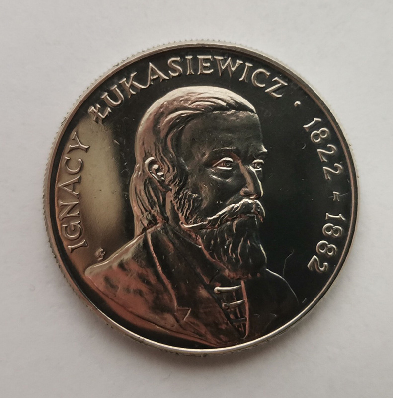 Srebrna moneta z wizerunkiem mężczyzny ukazanym w profilu. Mężczyzna ma długie włosy opadające na kark brodę i wąsy. Półkoliście nad postacią napis: Ignacy Łukasiewicz 1822-1882