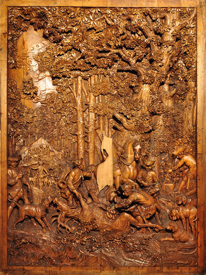 Autor nieznany, „Polowanie” płaskorzeźba; Niemcy(?), 2. połowa XIX wieku (?), 137 x 100,5 cm; relief, drewno lipowe Płaskorzeźba ma kształt pionowego prostokąta. Na pierwszym planie, nieco w prawo, myśliwy na koniu, po jego bokach łowczy i psy przy powalonym na grzbiet jeleniu z rozpłatanym brzuchem. Po bokach tej scenki jeszcze dwaj łowczy, ten z prawej stoi nad parującym kociołkiem. Na drugim planie, po lewej, namiot na polanie otoczonej półkolem niskich drzew. Przed namiotem ucztujący myśliwi z damą, poniżej namiotu łowczy z psem, za namiotem widoczny fragment czterokonnego zaprzęgu. W tle wysokie drzewa z dekoracyjnie potraktowanym listowiem. Postacie ubrane w stroje z XVIII wieku.