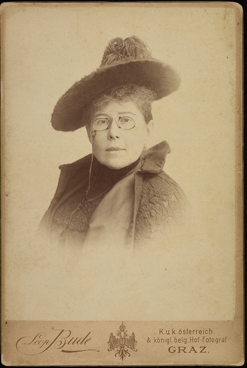 Maria Konopnicka, fotografia Leopolda Bude, z 1897 roku. Na fotografii popiersie kobiety w średnim wieku, w okularach, patrzącej na wprost. Ubrana jest w płaszcz ozdobiony gipiurą i kapelusz.