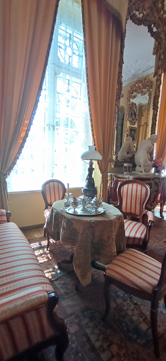 Fotografia przedstawia fragment wnętrza. Pośrodku stolik nakryty obrusem w jasnym kolorze z ciemniejszą dekoracją. Na stoliku komplet naczyń w kolorze srebrnym, na srebrnej tacy. Za nimi lichtarz ze świeczką i lampa z białym abażurem i ciemnym korpusem. Wokół stolika komplet mebli, z prawej strony trzy krzesła i z lewej fragment kanapy. Meble w ciemnobrązowym obramowaniu, z obiciem w pasy, jasne i czerwone na przemian, z delikatnym ornamentem. W tle okno z firanką i jasnobrązową kotarą z lambrekinem i frędzlami. Po prawej stronie lustro z ozdobną ramą. Pod lustrem szafka, na niej rzeźba w białym kolorze.