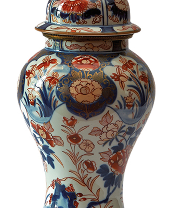 Fotografia kolorowa przedstawia fragment porcelanowego korpusu lampy naftowej. Na korpusie namalowane są stylizowane kwiaty lilii, goździków i chryzantem. Dekoracja w kolorystyce bieli, kobaltu, czerwieni ze złoceniami.