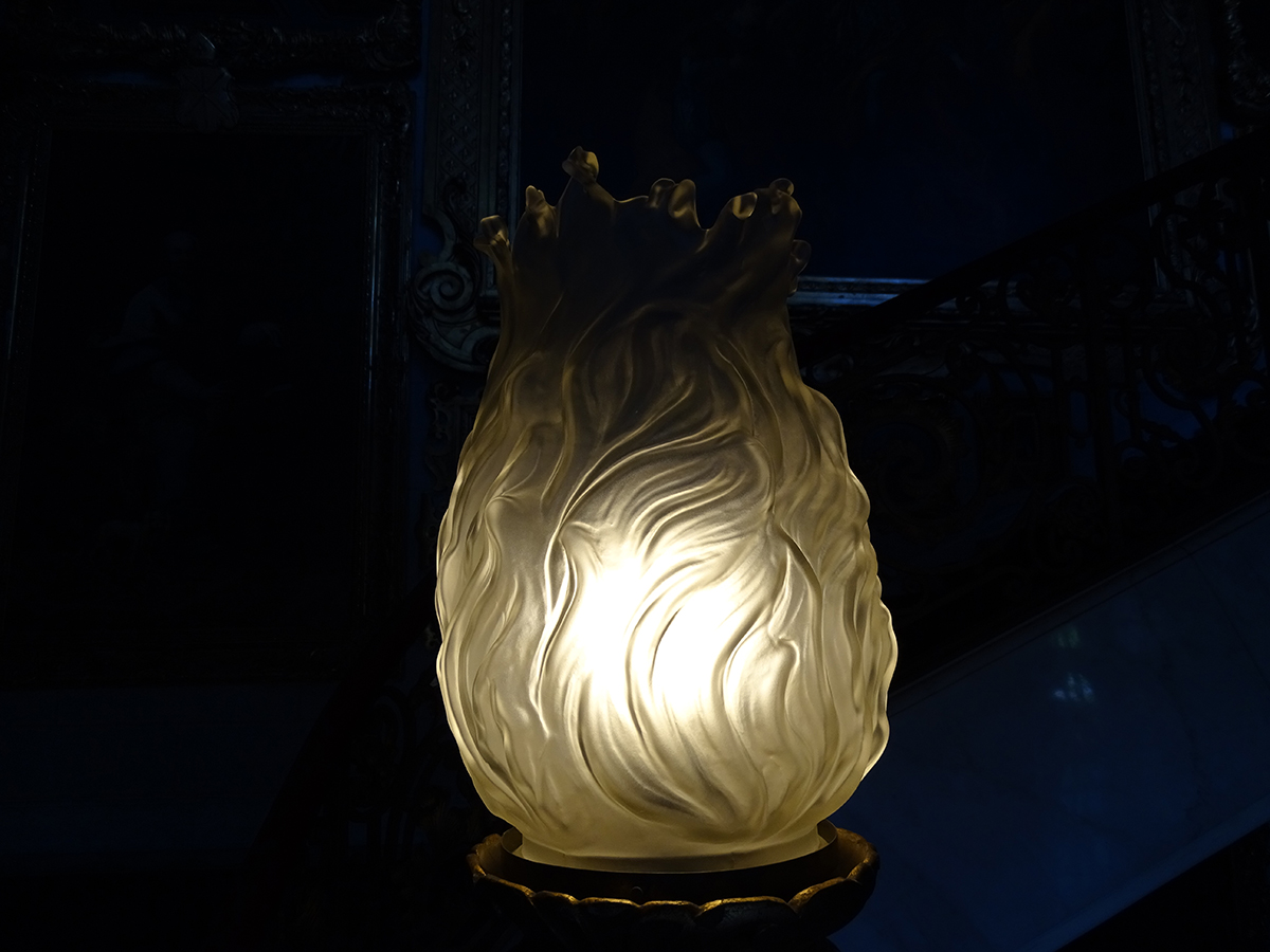 Fotografia kolorowa, współczesna przedstawia rozświetlony od wewnątrz szklany klosz lampy. W kształcie płomieni. Tło ciemne.