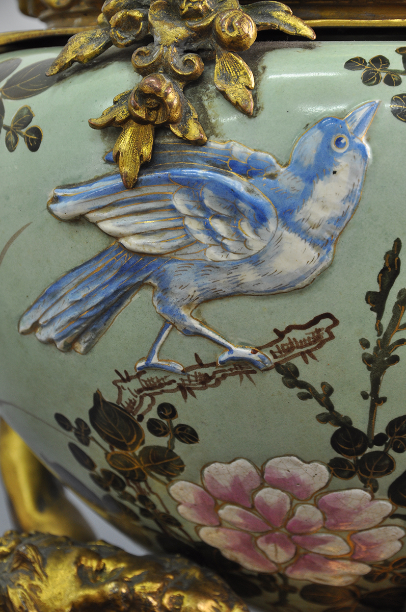 Fotografia kolorowa przedstawiająca fragment malarskiej dekoracji korpusu lampy naftowej. W centralnym miejscu widoczny jest półplastyczny biało-niebieski ptak siedzący na gałązce, poniżej różowy kwiat chryzantemy, powyżej fragment mosiężnej dekoracji. Tło w kolorze jasnozielonym.