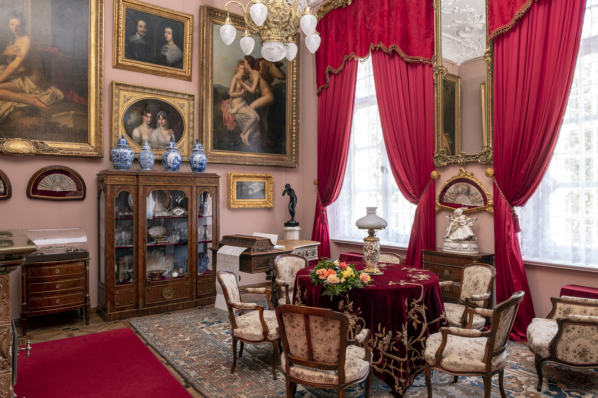 Współczesna kolorowa fotografia przedstawiająca wnętrze Małego Salonu w pałacu kozłowieckim. Po prawej w tle widoczne wysokie okna udekorowane czerwonymi lambrekinami i kotarami ze złoconą bordiurą, w oknach śnieżnobiałe firanki sięgające parapetu, pomiędzy oknami wysokie wąskie lustro kryształowe ze złoconą ramą. Poniżej w złoconej oprawie wachlarz i porcelanowa figura na szafce. Po prawej stronie w tle obrazy różnej wielkości oprawione w złocone ramy, poniżej przeszklona witryna z kryształami i srebrami. Na samym dole wśród drobiazgów pudełko po słodyczach z cukierni „Zjednoczeni Cukiernicy Warszawscy”. Na witrynie 4 fajansowe biało-kobaltowe wazy. W rogu pomieszczenia na postumencie ciemna figura kobiety pijącej z czarki. Na pierwszym planie stół okrągły przykryty mięsistą aksamitną tkaniną w kolorze ciemnego bordo, ze złotą wypukłą aplikacją. Na stole lampa naftowa o jasnej, porcelanowej podstawie i cebulastym białym kloszu. Wokół stołu fotele i krzesła tapicerowane w stylu Ludwika Filipa, za stołem przypominający kształtem fortepian włoski mechanizm pianomelodikon, odtwarzający muzykę z papierowych perforowanych taśm.