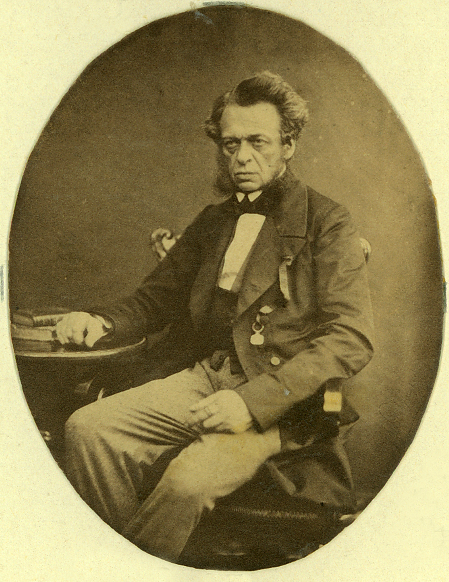 Fotografia archiwalna, w kolorze sepii, owalna, przedstawiająca starszego mężczyznę siedzącego przy stoliku, zwrócony en trois quarts w lewo; tło neutralne.