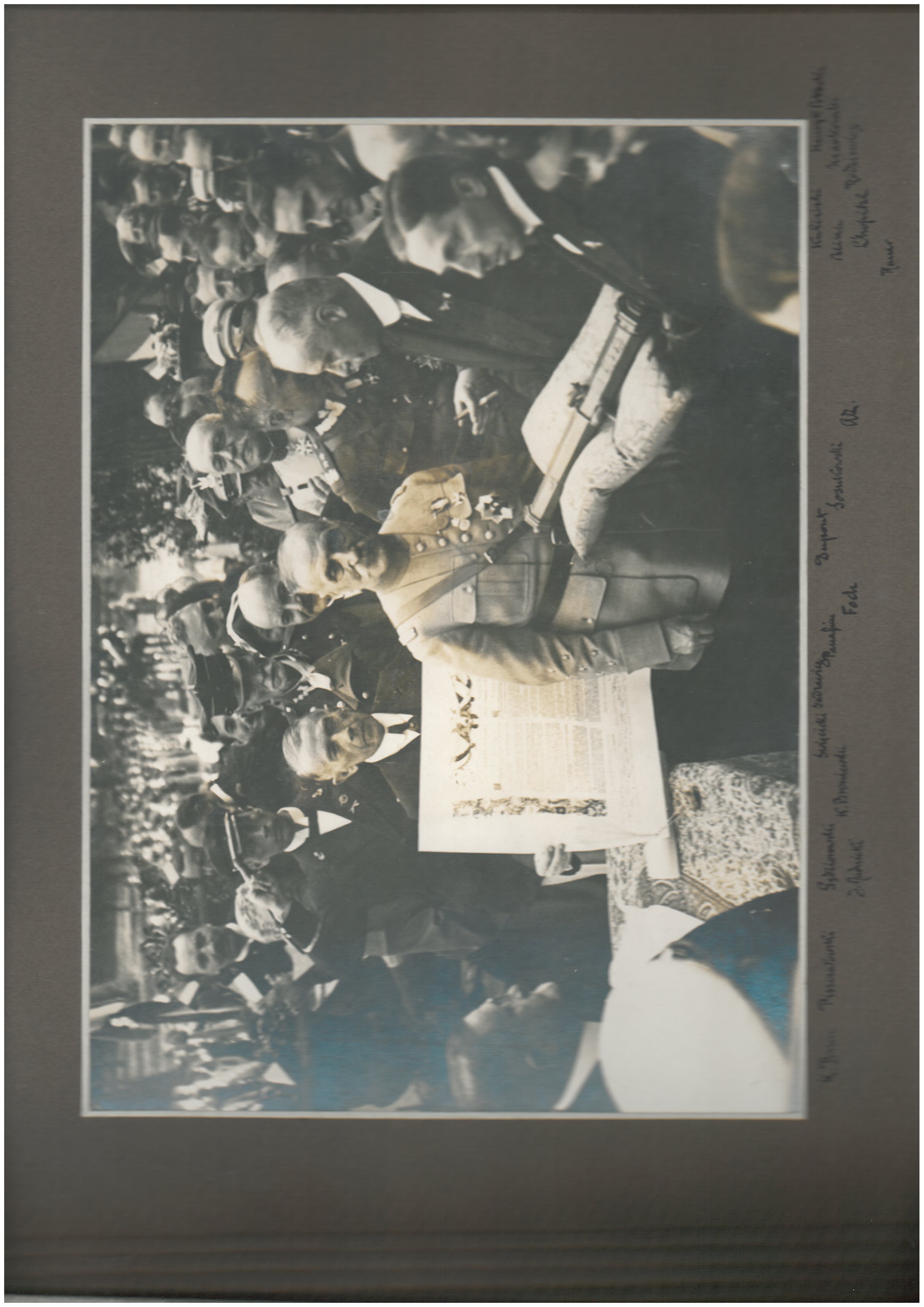 Zdjęcie jednej z kart w albumie pamiątkowym – na ciemnym tle naklejone zdjęcie archiwalne, czarno-białe, przedstawiające grupę mężczyzn w różnym wieku w . Pośrodku starszy mężczyzna w mundurze wojskowym z różnymi odznaczeniami na lewej piersi. Obok niego, nieco z przodu mężczyzna w garniturze trzymający na ozdobnej poduszce futerał; za nim mężczyzna trzymający ozdobny afisz z tekstem w języku polskim i francuskim.