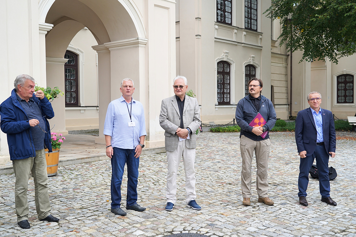 Oficjalne otwarcie Turnieju Drużyn Muzealnych Wewnętrznych Służb Ochrony Na zdjęciu widać pięciu mężczyzn stojących na dziedzińcu przed wejściem do pałacu. Wśród nich p. Andrzej Pruszkowski, Dyrektor Muzeum Zamoyskich w Kozłówce, który otworzył uroczystość.
Tło stanowi jasna, klasycystyczna fasada budynku z dużymi, ciemnymi oknami oraz arkadą. Na dziedzińcu ułożona jest kamienna kostka. W rogu kadru widać drewnianą donicę z kwiatami oraz fragment drzewa z zielonymi liśćmi.
Mężczyźni ustawieni są w lekkim półkolu, zwróceni w stronę fotografa:
pierwszy z lewej ma siwe włosy, ubrany jest w niebieską kurtkę i oliwkowe spodnie, trzyma w ręku okulary, obok stoi mężczyzna w jasnej koszuli z identyfikatorem na smyczy i dżinsach, na środku znajduje się starszy pan w jasnoszarej marynarce i białych spodniach, ze złożonymi rękami, czwarty mężczyzna ma ciemne włosy i wąsy, ubrany jest w szarą bluzę i jasne spodnie, trzyma w rękach teczkę w bordowym kolorze, po prawej stoi mężczyzna w granatowym garniturze i błękitnej koszuli, z rękami opuszczonymi wzdłuż ciała.
Cała grupa uczestniczy w oficjalnym otwarciu turniej WSO na dziedzińcu Muzeum.
