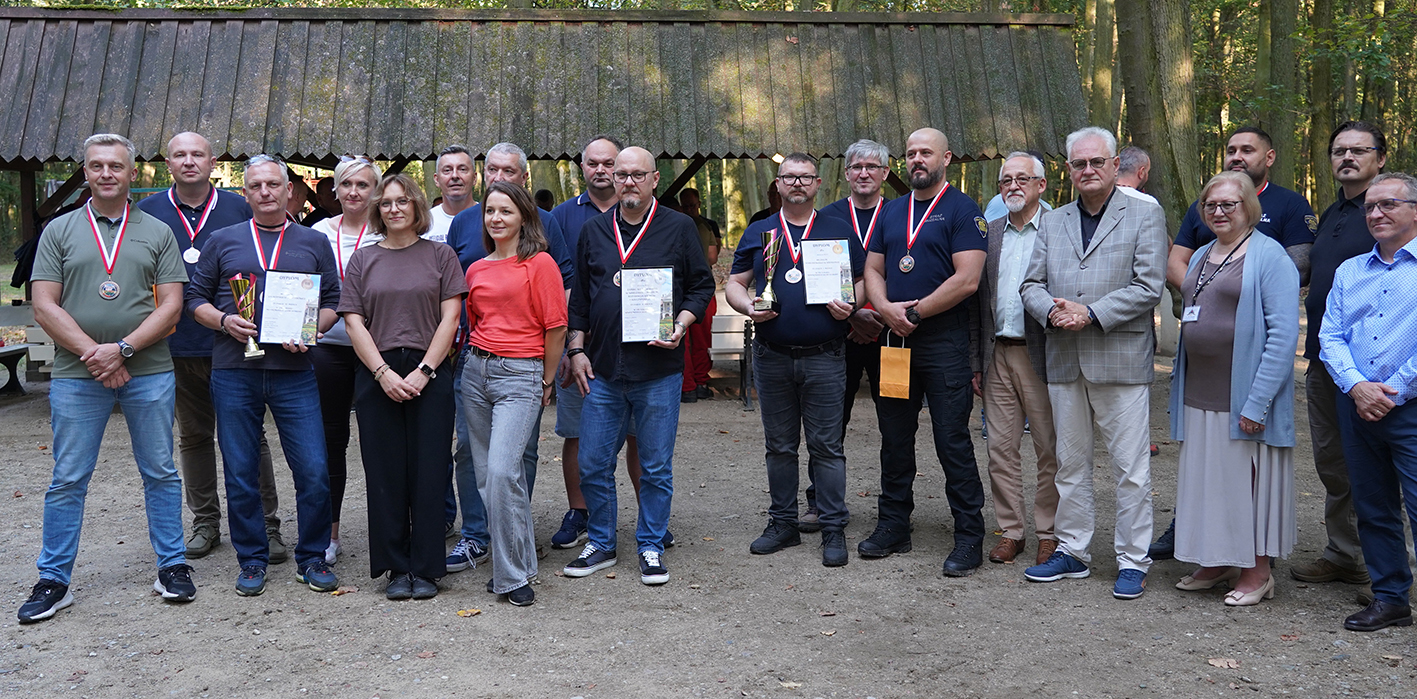 Zdjęcie grupowe na zakończenie Turnieju Na zdjęciu widoczna jest grupa kilkunastu osób stojących wspólnie w plenerze, na tle drewnianej wiaty i drzew w lesie. To fotografia pamiątkowa wykonana podczas wydarzenia lub uroczystości wręczenia nagród. Jest to podsumowanie turnieju WSO w Muzeum.
Na pierwszym planie stoją mężczyźni i kobiety w różnym wieku, część z nich ma na szyjach medale, a w rękach trzyma dyplomy i puchary – to prawdopodobnie laureaci zawodów lub konkursu. Widać m.in. trzy osoby z medalami i dyplomami oraz kilka osób w ciemnych koszulkach sportowych. Obok zwycięzców stoją inne osoby, prawdopodobnie organizatorzy i goście, w strojach bardziej formalnych: marynarkach, koszulach, a także eleganckich butach.
Wszyscy patrzą w stronę obiektywu, uśmiechając się lub zachowując poważniejszy wyraz twarzy. Tło stanowią drzewa i drewniana altana, co sugeruje, że uroczystość miała miejsce w leśnym otoczeniu, w nieformalnej, przyjaznej atmosferze.