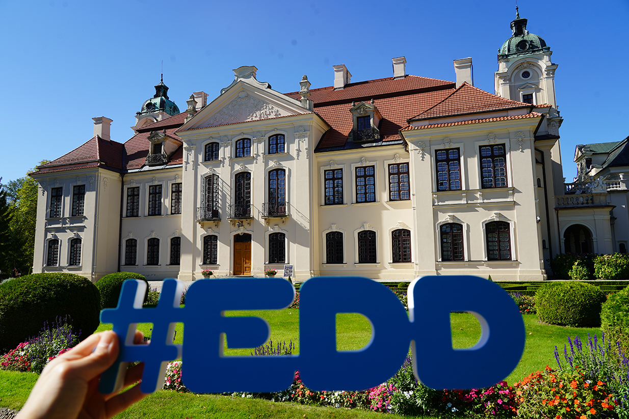 Europejskie Dni Dziedzictwa Na zdjęciu widać reprezentacyjny pałac w Kozłówce – siedzibę Muzeum Zamoyskich. Budynek ma jasną, kremową elewację, liczne wysokie okna w ciemnych ramach i czerwony dach kryty dachówką. Zwieńczony jest dekoracyjnymi szczytami oraz wieżyczkami z zielonymi kopułami.
Na pierwszym planie widzimy rękę trzymającą duży, niebieski napis w formie hasztagu: #EDD – nawiązujący do Europejskich Dni Dziedzictwa. W tle, przed pałacem, rozciąga się starannie utrzymany ogród z kolorowymi kwiatami i przyciętymi krzewami.
Całość fotografii, wykonanej w słoneczny dzień, oddaje elegancję rezydencji i podkreśla jej rolę jako miejsca wydarzeń kulturalnych.