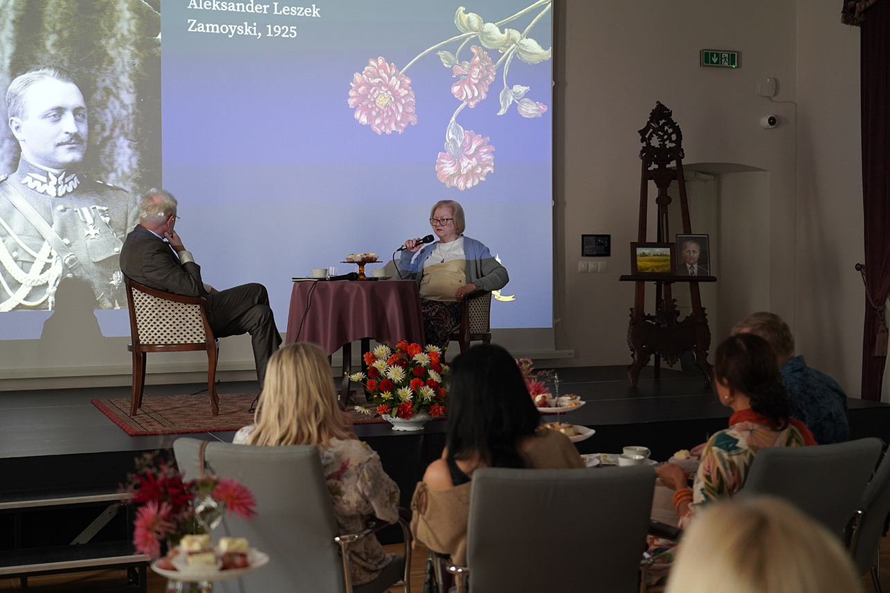 Herbatka z Hrabiną Na fotografii widzimy wydarzenie odbywające się w eleganckiej sali teatralnej Muzeum Zamoyskich w Kozłówce.
Na pierwszym planie, tyłem do obiektywu, siedzą uczestnicy spotkania przy stolikach nakrytych białą zastawą, z filiżankami herbaty oraz paterami z ciastem i kanapkami.
W centrum kadru znajduje się scena. Przy okrągłym stoliku nakrytym bordowym obrusem siedzą dwie osoby. Po lewej stronie, pan Andrzej Pruszkowski, Dyrektor Muzeum Zamoyskich, prowadzący to spotkanie. Mężczyzna siedzi bokiem do publiczności, z uwagą wsłuchując się w opowieść Inki. Po prawej, wspomniana Jadwiga Inka Zamoyska, ubrana w jasny sweter, w okularach, trzyma mikrofon opowiada historię.
Za rozmówcami widoczny jest ekran z dużą projekcją – czarno-białym portretem mężczyzny w mundurze z 1925 roku, podpisanym jako Aleksander Leszek Zamoyski. Jest to ostatni właściciel Kozłówki, tata Inki. Ekran ozdobiony jest także ilustracją kwiatów.
Na scenie, obok ekranu, stoi drewniana sztaluga, na której ustawiono ramki ze zdjęciami – jedno przedstawia portret mężczyzny, drugie pejzaż. Przed stolikiem ustawiono dużą, kolorową kompozycję kwiatową w odcieniach czerwieni, bieli i pomarańczu.