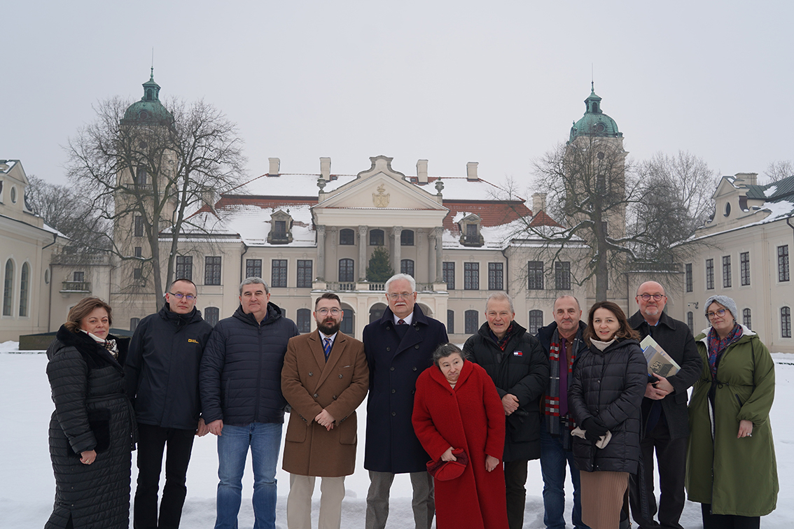 Na zdjęciu widoczna jest grupowa fotografia plenerowa wykonana zimą. Kadr jest szeroki, a tło stanowi jasny, symetryczny zespół pałacowy: w centrum znajduje się reprezentacyjny budynek z portykiem i trójkątnym frontonem, po bokach widać dwie wysokie wieże zakończone kopułami w zielonkawym kolorze. Dachy i parapety są przysypane śniegiem, a po obu stronach stoją bezlistne drzewa. Niebo jest jasnoszare i pochmurne.
Na pierwszym planie, na śniegu, stoi jedenaście osób ustawionych w jednym rzędzie, zwróconych przodem do aparatu. Większość ma na sobie zimowe, ciemne okrycia.
Od lewej strony:
1.	Kobieta w długim, czarnym pikowanym płaszczu i szaliku.
2.	Mężczyzna w ciemnej kurtce z wysokim kołnierzem, w okularach.
3.	Mężczyzna w granatowej, pikowanej kurtce.
4.	Mężczyzna z brodą i okularami, w jasnobrązowym, eleganckim płaszczu.
5.	W centrum starszy mężczyzna w długim, ciemnogranatowym płaszczu; stoi wyprostowany.
6.	Przed nim i nieco niżej stoi kobieta w intensywnie czerwonym płaszczu, trzyma w dłoniach czerwone nakrycie głowy.
7.	Mężczyzna w czarnej kurtce.
8.	Mężczyzna w ciemnej kurtce, z szalikiem w czerwono-granatowe pasy.
9.	Kobieta w czarnej, pikowanej kurtce i jasnym szaliku.
10.	Mężczyzna w czarnym płaszczu, w okularach; pod pachą trzyma teczkę lub książkę.
11.	Kobieta w zielonym płaszczu, w czapce i szaliku.
Całość ma charakter oficjalnego pamiątkowego zdjęcia: postacie stoją blisko siebie, spokojnie, bez wyraźnych gestów, na tle zabytkowej architektury i zimowego krajobrazu.