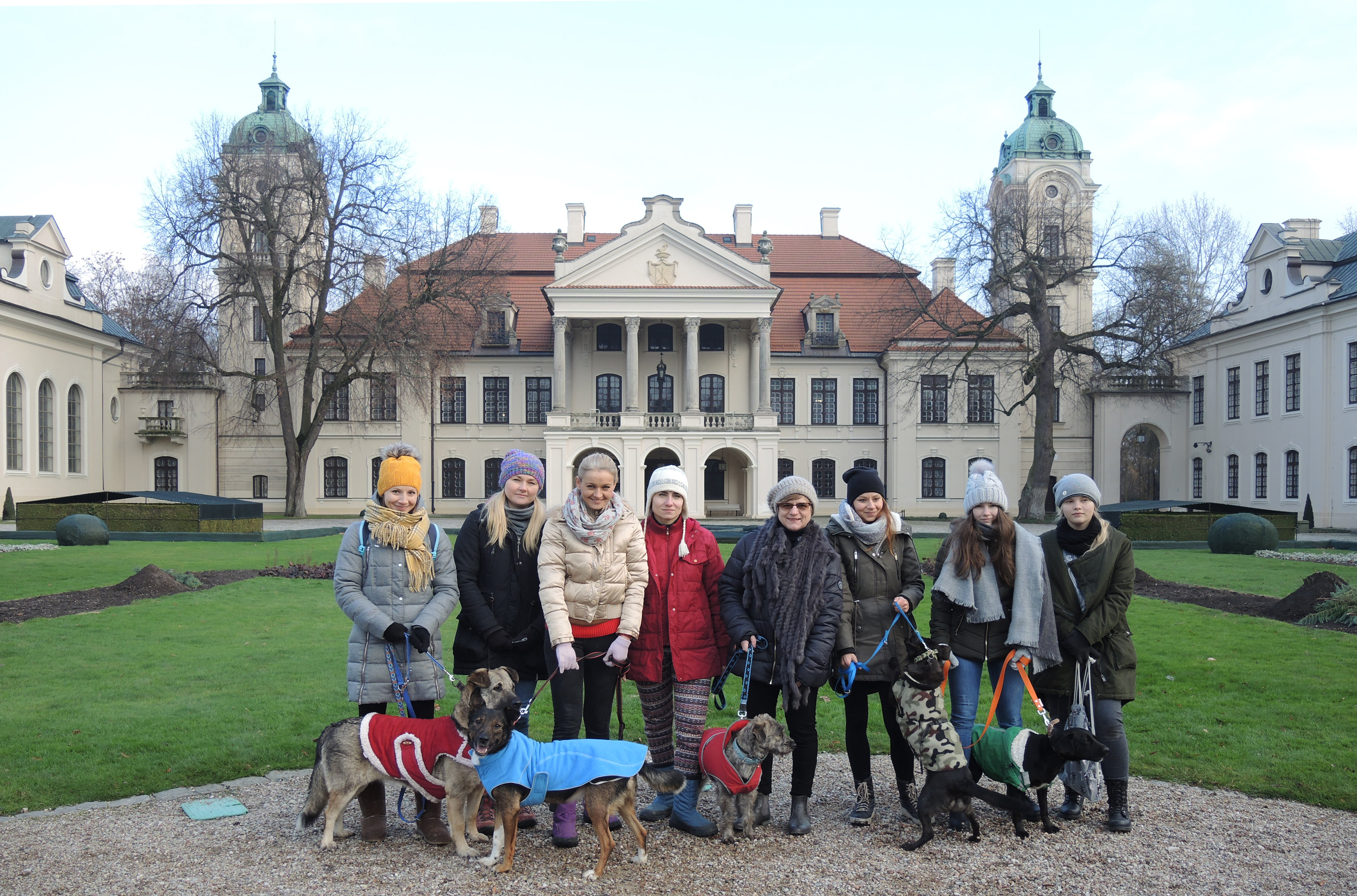Fotografia. Grupa ośmiu kobiet w różnym wieku, ubrana w zimowe kurtki i czapki stojąca na tle okazałej dwukondygnacyjnej rezydencji z portykiem i czerwonym dwuspadowym dachem oraz pobocznymi budynkami tworzącymi literę „U”. Pięć z nich trzyma psy na smyczy.