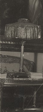 Fotografia archiwalna czarno biała fotografia przedstawiająca lampę ustawioną na stole we wnętrzu pomieszczenia. Lampa ma kolumnową podstawę i tekstylny szeroki i płaski  abażur.