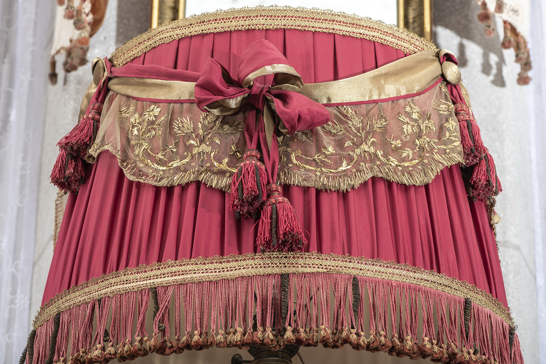 Fragment abażuru lampy naftowej dekorowany oryginalną pasmanterią, Klatka Schodowa pałacu kozłowieckiego; zbiory Muzeum Zamoyskich w Kozłówce, nr inw. MPK/T/83 Fotografia przedstawia fragment abażuru z czerwonej tkaniny, udekorowanej materiałową szarfą w kolorze złota w połączeniu z tiulem wyszywanym wzorem kwiatowym.