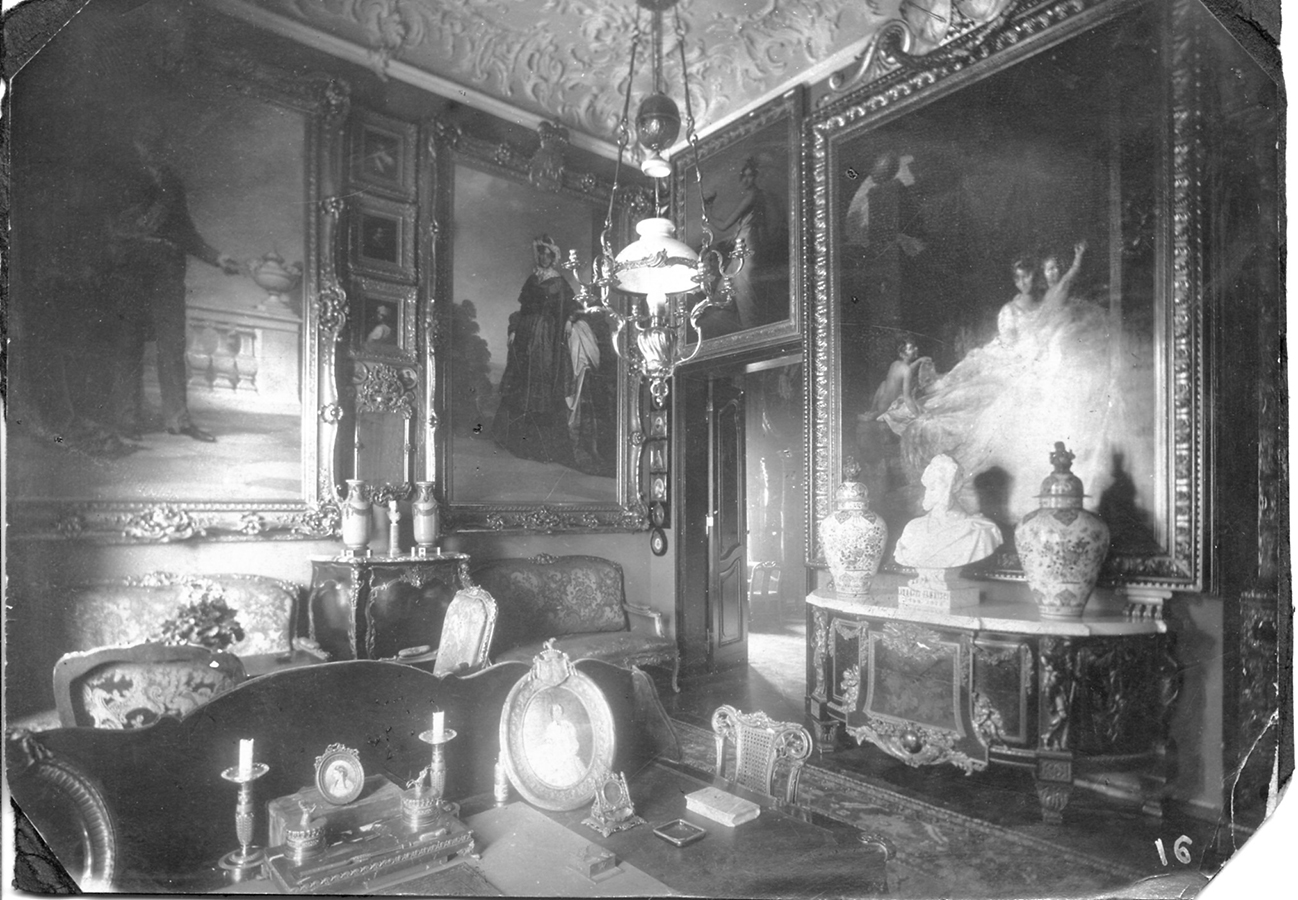 Gabinet Konstantego Zamoyskiego, około 1900 roku; fotografia archiwalna; nr inw. MPK/F/1898 Czarno-biała fotografia archiwalna przedstawiająca wnętrze Gabinetu hrabiego Konstantego Zamoyskiego około 1900 roku. Na pierwszym planie widoczny fragment biurka, na nim przybory do pisania i oprawione w stylowe ramki wizerunki matki i żony hrabiego. W głębi za biurkiem stylowe meble, po prawej stronie bogato zdobiona okazała komoda. Na niej białe marmurowe popiersie i dwie wazy. Obok widoczne otwarte drzwi prowadzące do kolejnego pomieszczenia. Na ścianach gęsto zawieszone różnej wielkości obrazy oprawione w dekoracyjne ramy. Z sufitu zwisa lampa naftowo-świecznikowa na dekoracyjnych łańcuchach.