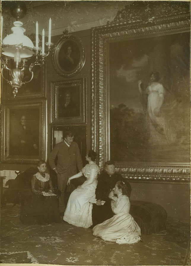 Konstanty i Aniela Zamoyscy w towarzystwie siostrzenic i nieznanej damy w północnej sypialni pałacu kozłowieckiego; fotografia archiwalna; nr inw. MPK/F/38 Fotografia archiwalna w sepii przedstawiająca pięć osób, Konstantego i Anielę Zamoyskich w towarzystwie siostrzenic i nieznanej damy w północnej sypialni pałacu kozłowieckiego. Pomieszczenie udekorowane różnej wielkości obrazami, oprawionymi w złocone i bogato zdobione ramy. Mężczyzna w garniturze stoi zwrócony w stronę młodej kobiety w jasnej sukni z ciemnymi, wysoko upiętymi włosami, siedzącej na szezlongu. Obok niej siedzi druga kobieta, starsza, w czarnej sukni. Przy niej siedząca na dywanie druga młoda kobieta w jasnej sukni. Po lewej stronie mężczyzny siedzi uśmiechnięta kobieta w średnim wieku w ciemnej sukni z kołnierzem. Na jej kolanach widoczny jest dokument. W lewym górnym rogu świecznikowo-naftowa lampa wisząca.