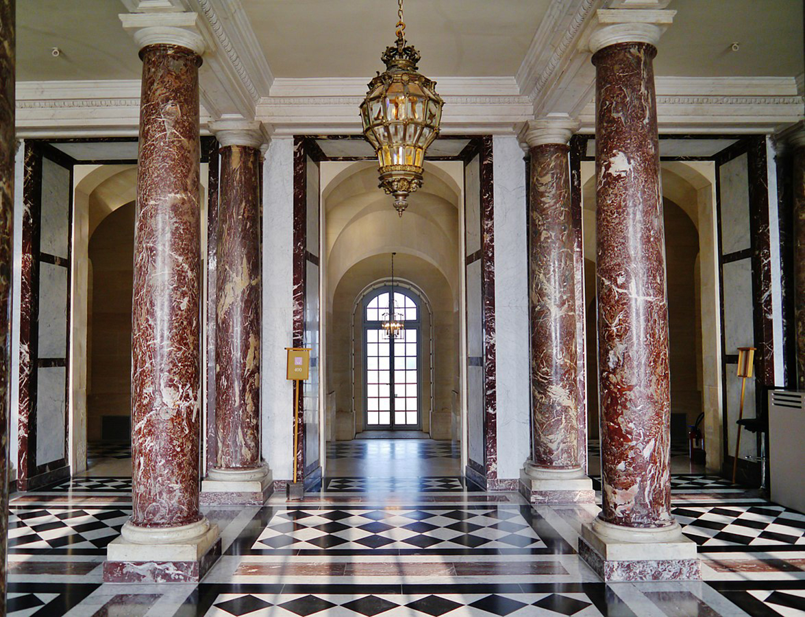 Westybul Marmurowy w Wersalu, stan obecny; autor: Zairon, CC BY-SA 4.0 <https://creativecommons.org/licenses/by-sa/4.0>, via Wikimedia Commons, źródło: File: Versailles Château de Versailles Appartements d'Hiver 01.jpg - Wikimedia Commons, dostęp 18.05.2022 Fotografia kolorowa, współczesna, przedstawiająca wnętrze Westybulu Marmurowego w pałacu w Wersalu. Jasne pomieszczenie z czterema marmurowymi kolumnami w odcieniach brązu i wysokimi przejściami. W głębi, pośrodku widoczne zakończone półkoliście okno podzielone listwami. Przy suficie złocona latarnia w kształcie odwróconej gruszki z przeszkleniem w postaci kwadratowych szybek.