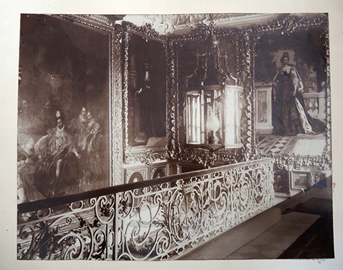 Klatka schodowa pałacu kozłowieckiego, po 1900 roku; fotografia archiwalna; nr inw. MPK/F/1881 Archiwalne zdjęcie w sepii, ukazujące piętro reprezentacyjnej klatki schodowej pałacu w Kozłówce, około 1900 roku. Wzdłuż posadzki zamocowana jest balustrada dekorowana liśćmi akantu i pośrodku herbem rodziny Zamoyskich, Jelita, przedstawiającym trzy skrzyżowane kopie, zwieńczonym dziewięciopałkową koroną hrabiowską. Poręcz wyłożona materiałem. Nad balustradą widoczna duża wisząca latarnia o metalowej konstrukcji i kryształowych taflach. Wewnątrz jasny szklany klosz. W tle na ścianach obrazy oprawione w dekorowane, złocone ramy.