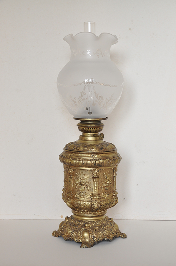 Lampa naftowa z dekoracją kwiatową, XIX wiek; stop metali, mosiądz, szkło; nr inw. MPK/ZM/1405 Fotografia kolorowa przedstawia lampę w formie przysadzistego pucharu pokrytego ornamentami, z tulipanowym kloszem o wyniesionej falistej krawędzi. Na walcowatym korpusie architektoniczna dekoracja z półplastycznych pilastrów, pomiędzy nimi wazony z bukietami kwiatów. Klosz w formie spłaszczonej kuli z dość wysoką kryzą, dekorowany trawionymi stylizowanymi girlandami, połączonymi odcinkami łańcucha.