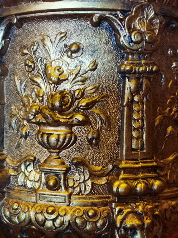Fragment dekoracji korpusu lampy naftowej z dekoracją kwiatową, XIX wiek; stop metali, mosiądz, szkło; nr inw. MPK/ZM/1405 Fotografia kolorowa przedstawia wypukłą dekorację mosiężnego korpusu lampy złożoną z półplastycznych pilastrów, między którymi umieszczone są wazony z bukietami kwiatów.