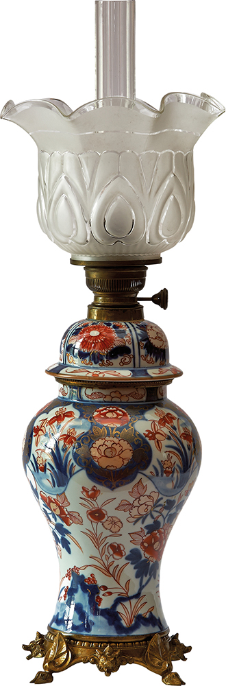 Lampa naftowa; korpus: Arita, Japonia, porcelana; palnik: Ehrich und Greatz, Berlin, Niemcy, mosiądz; 2. połowa XIX wieku; nr inw. MPK/CS/458 Fotografia kolorowa przedstawia lampę naftową o porcelanowym, wybrzuszonym korpusie osadzonym na mosiężnej podstawie wspartej na czterech nóżkach. Na korpusie namalowane są stylizowane kwiaty lilii, goździków i chryzantem. Dekoracja w kolorystyce bieli, kobaltu, czerwieni ze złoceniami. Klosz lampy ma tulipanowy kształt.