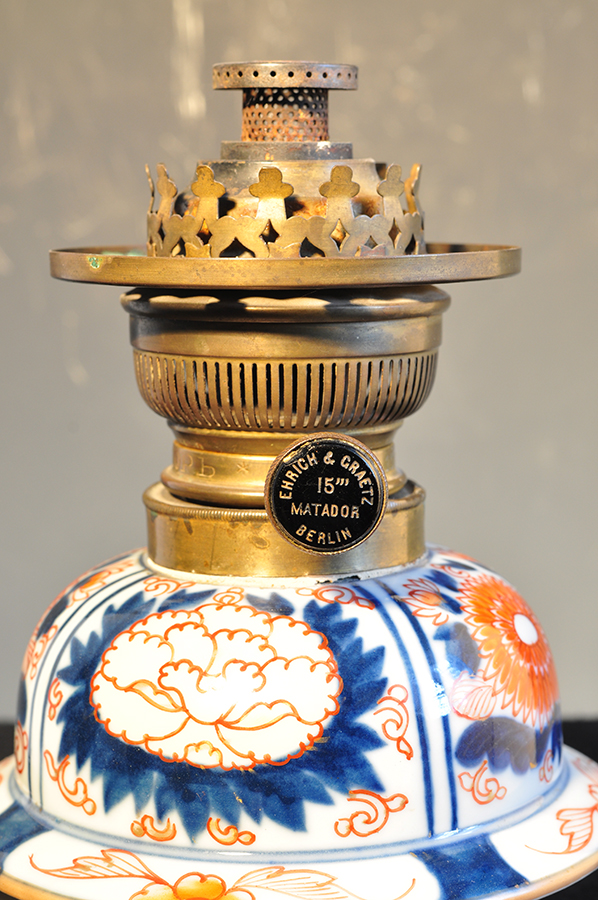 Lampa naftowa; korpus: Arita, Japonia, porcelana; palnik: Ehrich und Greatz, Berlin, Niemcy, mosiądz; 2. połowa XIX wieku; nr inw. MPK/CS/458 Fotografia kolorowa przedstawia fragment lampy naftowej. Widoczna część korpusu porcelanowego ozdobiona jest wzorem kwiatowym w kolorach białym, kobaltowym, czerwonym ze złoceniami. Na niej osadzony jest palnik, który w niższej części mechanizmu ma zamocowane pokrętło do regulacji wysokości płomienia, wyżej perforowane elementy konstrukcji dostarczające tlenu. Całość palnika zakończona dekoracyjną galeryjką. Na pinezce pokrętła, na czarnym tle złotym kolorem napis: półkoliście Ehrich und Greatz, Berlin – nazwa producenta i w centralnej części 15’’’ Matador – informacja o szerokości knota.