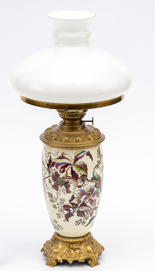 Lampa naftowa stołowa, salonowa; Anglia, początek XX wieku; mosiądz, fajans, szkło bezbarwne; nr inw. MPK/CS/2054 Fotografia kolorowa przedstawia lampę naftową o metalowej podstawie, na której osadzony jest owoidalny porcelanowy korpus ozdobiony kwiatami katlei i liściastymi gałązkami. Kolorystyka w odcieniach różu, błękitu i żółci. Klosz kopulasty, ze szkła mlecznego, współczesny.
