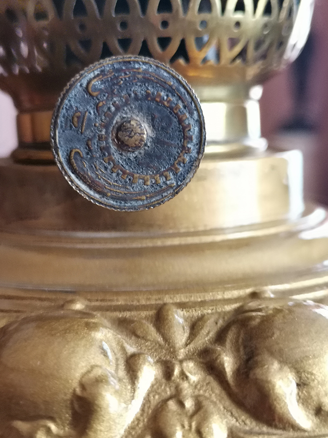 Fragment zbiornika i pokrętła lampy naftowej; Anglia, początek XX wieku; mosiądz; nr inw. MPK/CS/2054 Fotografia kolorowa przedstawia fragment mosiężnego zbiornika lampy naftowej oraz fragment palnika z pinezką pokrętła. Na pokrętle widoczny jest zatarty wzór producenta.