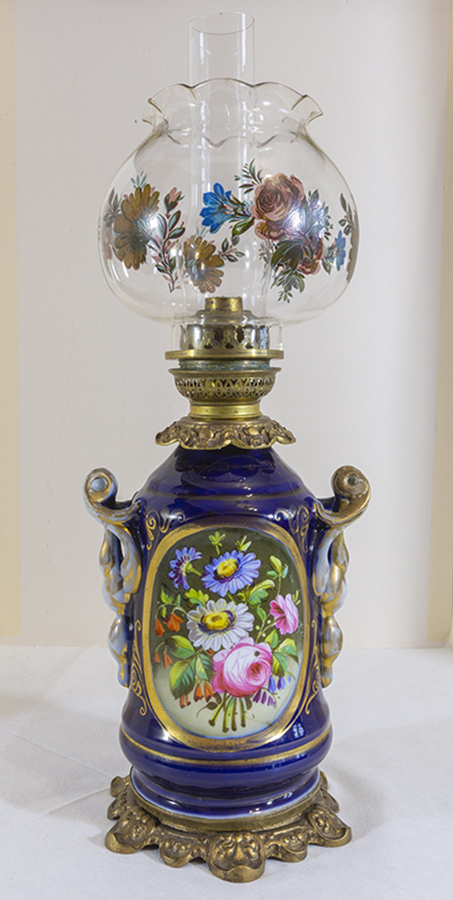 Lampa naftowa, Francja, Bayeaux – Valentine (?), 2. połowa XIX wieku; porcelana, szkło, farby naszkliwne, mosiądz; nr inw. MPK/CS/2212 Fotografia kolorowa przedstawia lampę naftową o porcelanowym kobaltowym korpusie z bukietami kwiatów w rewersach. W owalu bukiet kwiatów w kolorach białym, różowym, niebieskim, pomarańczowym. Pomiędzy kwiatami zielone liście. Podstawa mosiężna, klosz kulisty szklany, malowany w kwiaty.