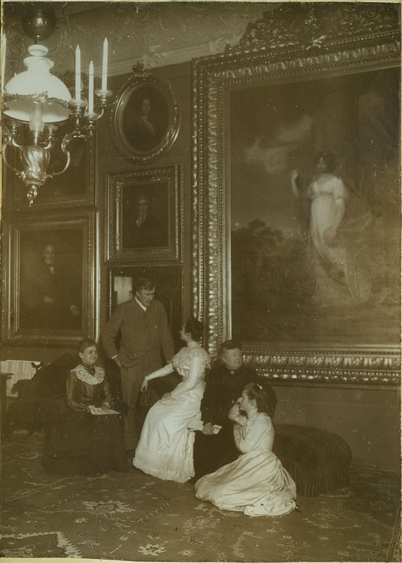 14. Wnętrze Sypialni Hrabiego Konstantego, Polska, początek XX wieku; fotografia zabytkowa; zbiory Muzeum Zamoyskich w Kozłówce, nr inw. MPK/F/38 Fotografia zabytkowa w kolorze sepii ukazuje kilka postaci we wnętrzu sypialni pałacu w Kozłówce. Od lewej: nieznana kobieta, na tle drzwi stoi Konstanty Zamoyski, obok siedzi nieznana kobieta, dalej Aniela z Potockich Zamoyska i klęcząca Zofia Wodzicka. W tle ściana z portretami. Największy przedstawia Zofię z Czartoryskich Zamoyską. W lewym górnym rogu fotografii widoczna lampa wisząca naftowo-świecowa.