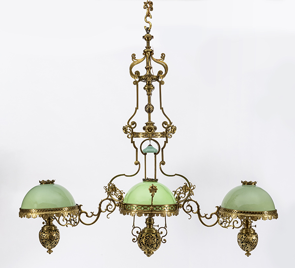 7. Potrójna, wisząca lampa naftowa, Warszawa (?), 2.połowa XX wieku; mosiądz, szkło; palnik: Towarzystwo Akcyjne Fabryki Lamp : Bracia Brünner, Hugo Schneider i R. Ditmar; nr inw. MPK/ZM/87 Fotografia kolorowa przedstawia na białym tle potrójną lampę naftową. Mosiężna konstrukcja o esowato wygiętych ramionach łączy trzy zbiorniki na naftę ukryte w ażurowych czaszach o stylizowanych motywach kwiatowych. Nad każdym ze zbiorników szklany klosz w zielonym kolorze.