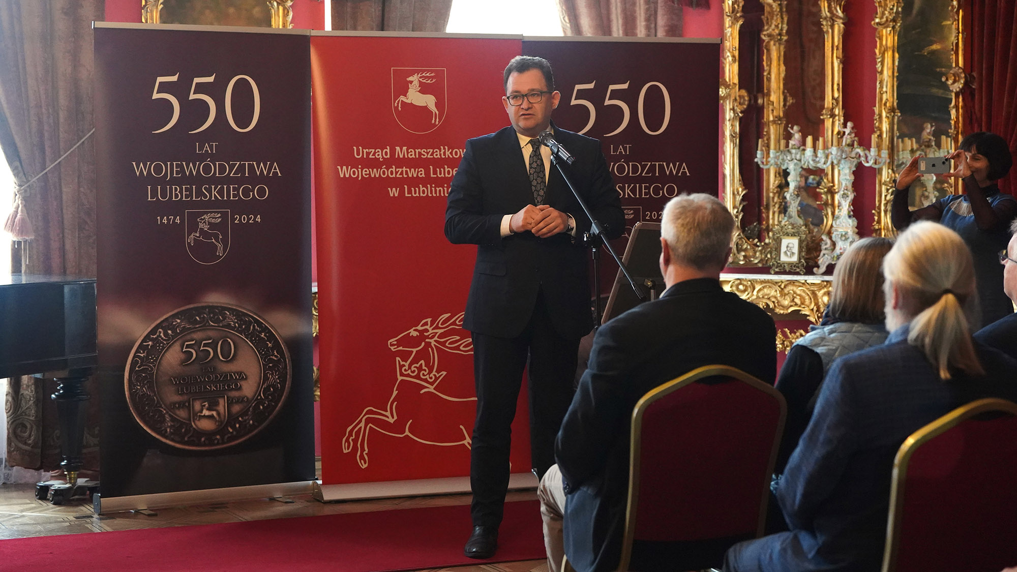 Członek Zarządu Województwa Lubelskiego, pan Bartłomiej Bałaban w trakcie przemowy. Mężczyzna ubrany w elegancki garnitur, przemawia trzymając dłonie złożone na piersi. Mówi do mikrofonu. W tle widoczne są elementy wystroju pałacowego, tworzące godne tła dla tej uroczystej chwili.