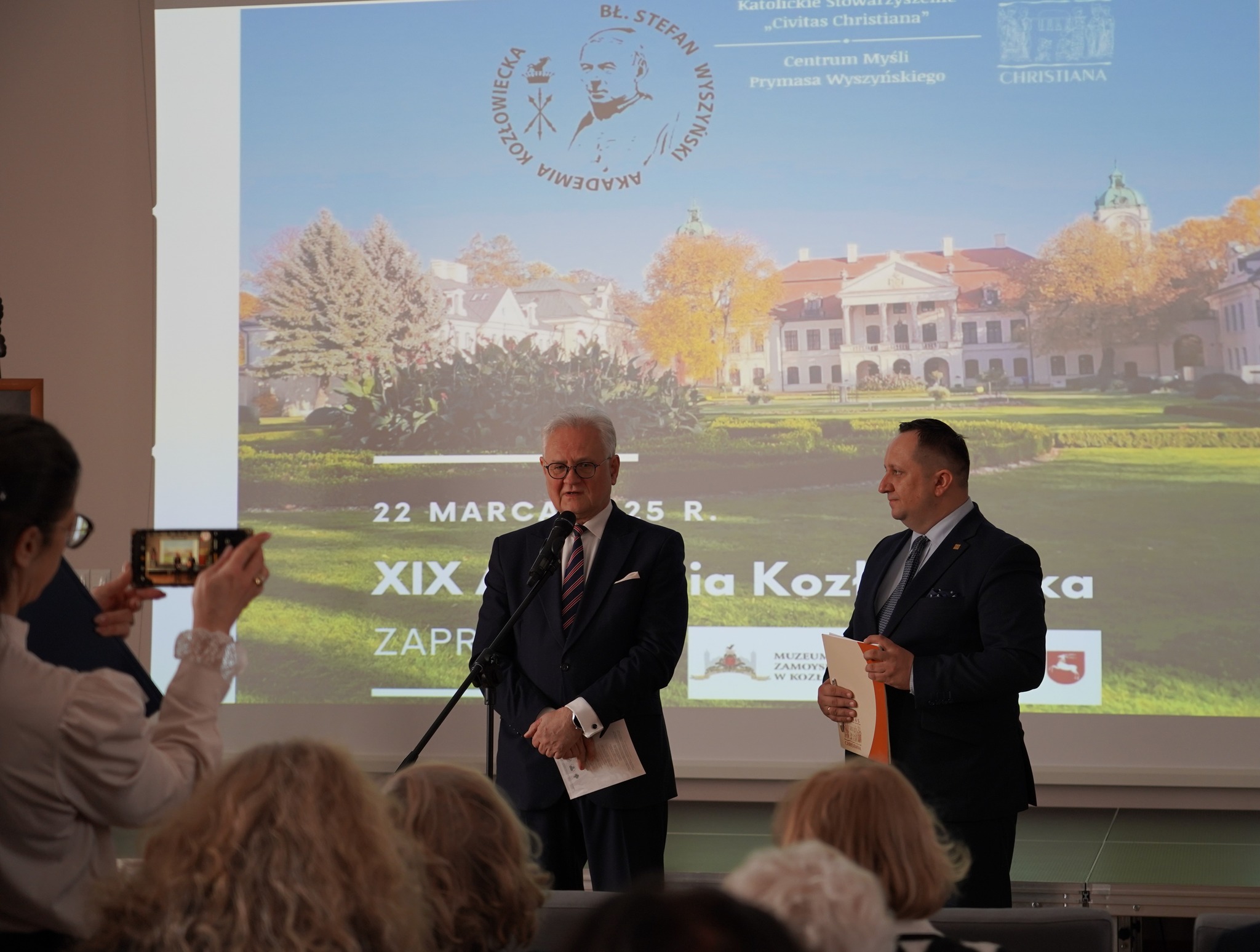 XIX konferencja Akademii Kozłowieckiej Na zdjęciu widać oficjalne wydarzenie odbywające się w zamkniętym pomieszczeniu, jest to XIX Akademia Kozłowiecka. Na pierwszym planie dwóch mężczyzn w eleganckich garniturach stoi przed mikrofonem, Andrzej Pruszkowski, p.o. Dyrektora Muzeum Zamoyskich oraz Marcin Sułek Członka Zarządu Stowarzyszenia Civitas Christiana. Starszy mężczyzna po lewej stronie, z siwymi włosami i okularami, mówi do mikrofonu, trzymając w dłoni kartkę papieru. Obok niego młodszy mężczyzna w ciemnym garniturze i krawacie słucha uważnie, trzymając w ręku dokumenty.
W tle znajduje się duży ekran, na którym wyświetlony jest slajd z napisem: "22 marca 2025 r. XIX Akademia Kozłowiecka" oraz grafiką przedstawiającą pałac otoczony zielonym ogrodem. Na ekranie widnieją także logotypy organizatorów wydarzenia, w tym Katolickiego Stowarzyszenia "Civitas Christiana" oraz Centrum Myśli Prymasa Wyszyńskiego.
Na lewym skraju zdjęcia kobieta w okularach i jasnej bluzce robi zdjęcie smartfonem, dokumentując wydarzenie. W dolnej części obrazu widać zgromadzoną publiczność – kilka osób siedzących w pierwszych rzędach, w tym osoby starsze o siwych włosach.
Atmosfera wydarzenia jest oficjalna i podniosła, a dobrze oświetlona sala i starannie dobrane stroje uczestników podkreślają formalny charakter spotkania.