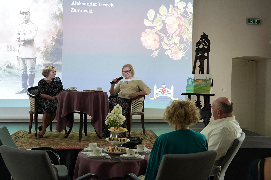 Podsumowanie spotkania “Herbatka z Hrabiną Jadwigą Inką Zamoyską” W eleganckiej, nastrojowo oświetlonej sali odbywa się spotkanie „Herbatka z Hrabiną Inką Zamoyską”. Na pierwszym planie widzimy gości siedzących przy okrągłych stolikach, nakrytych ciemnymi obrusami. Na stołach stoją filiżanki, imbryki, paterki z ciastkami i kwiatowe dekoracje. Goście słuchają z zainteresowaniem tego, co dzieje się na niewielkiej scenie.
Na scenie po prawej stronie siedzi kobieta z jasnymi włosami i okularami – to Hrabina Inka Zamoyska. Ubrana w jasny sweter i wzorzystą spódnicę, z uśmiechem trzyma mikrofon i dzieli się opowieścią. Obok niej, za ciemnym stolikiem, siedzi druga kobieta, prowadząca spotkanie, Pani Grażyna Antoniuk, również uśmiechnięta – prawdopodobnie moderatorka spotkania.
Za sceną wyświetlone jest czarno-białe archiwalne zdjęcie przedstawiające młodą parę w mundurach. Obok sceny stoją wysokie, stylowe świeczniki z zapalonymi sztucznymi świecami, dodające wnętrzu klimatu pałacowego salonu.
W tle widać wysokie okna przysłonięte ciężkimi, bordowymi zasłonami, przez które wpada rozproszone światło dzienne.