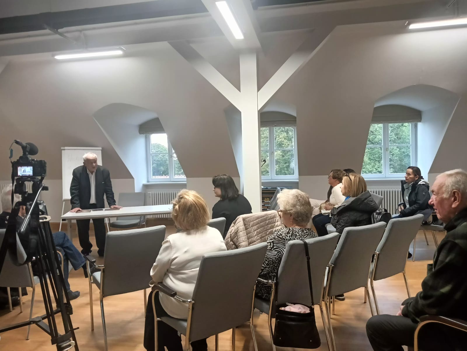 Zdjęcie przedstawia grupę kilku osób występujących w kameralnym wnętrzu o jasnych ścianach i czarno-białej, wzorzystej posadzce. Po lewej stronie kadru mężczyzna w okularach gra na akordeonie, siedząc na krześle. Obok niego, wzdłuż ściany i na schodach, stoi pięć osób – trzy kobiety i dwóch mężczyzn – śpiewających z nut w czarnych teczkach.
Wszyscy ubrani są elegancko, w stonowanych kolorach: panowie w ciemnych marynarkach, panie w jasnych bluzkach i spódnicach. Chór stoi na tle drewnianych schodów z ozdobną balustradą, na których ustawiono kilka sztalug z czarno-białymi grafikami.

Światło jest ciepłe i miękkie, podkreślające atmosferę skupienia i kameralności. Scena prawdopodobnie przedstawia występ chóru podczas finisażu wystawy artystycznej w muzeum.