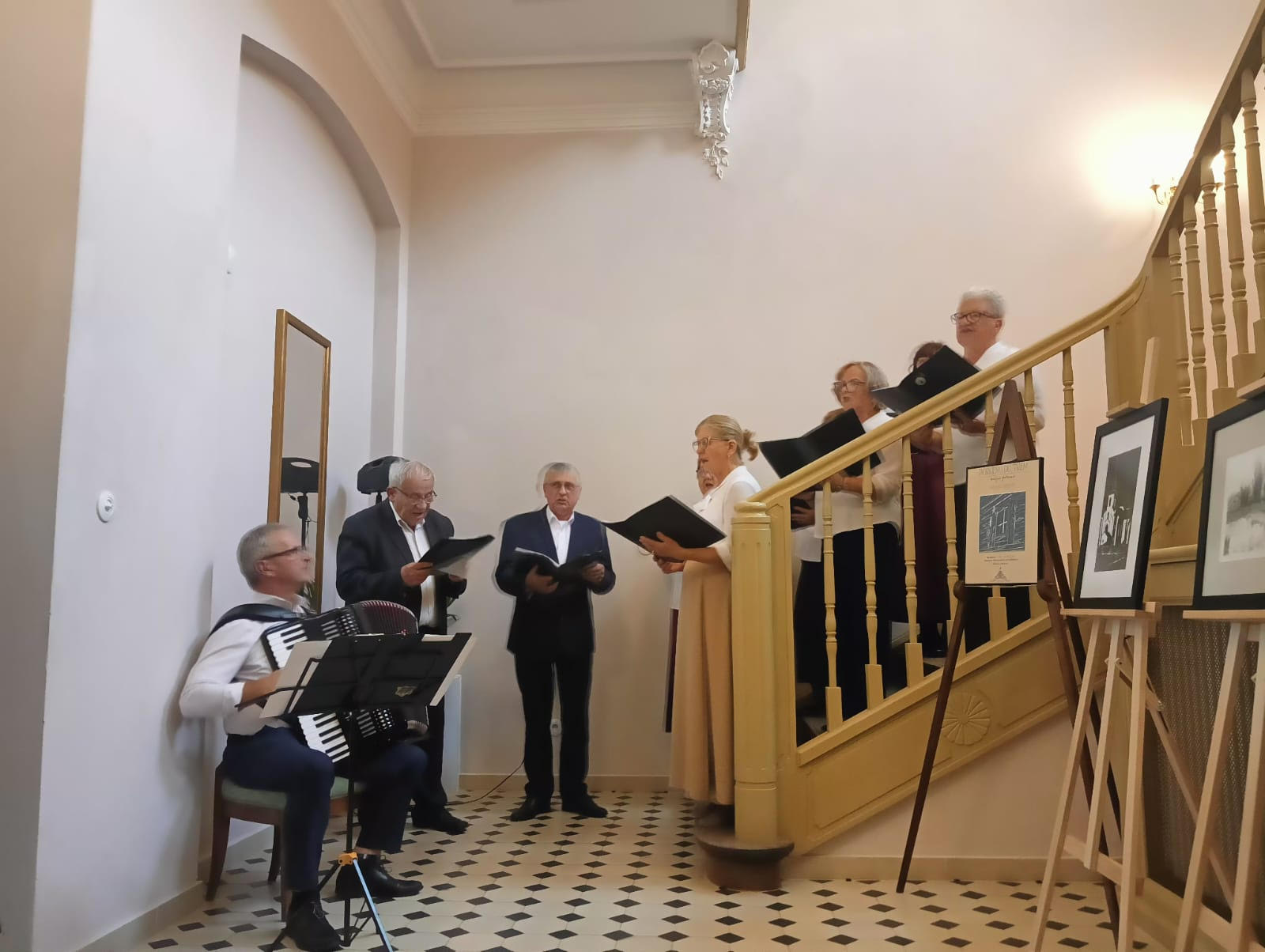 Zdjęcie przedstawia spotkanie w jasnej sali z ukośnym sufitem i dużymi oknami, przez które widać zieleń drzew. Na pierwszym planie po lewej stronie widać kamerę ustawioną na statywie, nagrywającą wydarzenie.
Przy stole na środku sali stoi starszy mężczyzna w ciemnej marynarce i białej koszuli, pochylony lekko do przodu, jakby prowadził wykład lub rozmowę. Przed nim siedzi grupa kilkunastu osób – głównie kobiet i kilku mężczyzn – uważnie słuchających. Uczestnicy zajmują miejsca w szarych, tapicerowanych krzesłach ustawionych w półkolu.
Atmosfera jest spokojna i skupiona. Światło dzienne wpadające przez okna podkreśla charakter edukacyjny i kameralny spotkania, prawdopodobnie o charakterze kulturalnym lub artystycznym.