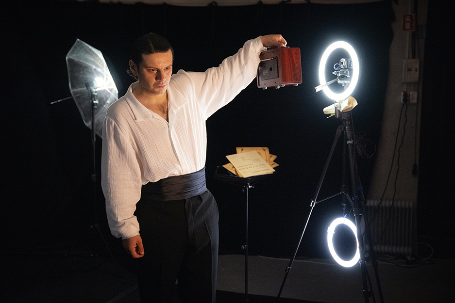 Zdjęcie przedstawia mężczyznę stojącego w ciemnej przestrzeni przypominającej scenę. Ma na sobie białą koszulę i ciemne spodnie. Jedną rękę unosi do góry, trzymając niewielkie urządzenie, drugą opuszcza wzdłuż ciała.

Obok niego znajduje się statyw z dwoma świecącymi okrągłymi lampami. W tle widoczne są elementy oświetlenia scenicznego oraz pulpit z kartkami.
