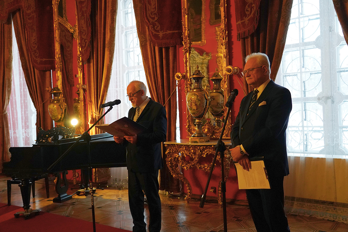 Zdjęcie przedstawia eleganckie, zabytkowe wnętrze pałacowe o bogatym wystroju. Dominują intensywne czerwienie ścian i ciężkie, bordowe zasłony z dekoracyjnymi lambrekinami, sięgające od sufitu do podłogi. W tle widoczne są wysokie okna, przez które wpada rozproszone światło dzienne, oraz duże, złocone lustra i ozdobne porcelanowe wazony ustawione na rzeźbionych konsolach.
Na pierwszym planie stoją dwaj starsi mężczyźni ubrani w ciemne, eleganckie garnitury. Obaj znajdują się przy mikrofonach na statywach. Mężczyzna po lewej stronie czyta tekst z kartki lub teczki, lekko pochylony w stronę mikrofonu. Obok niego, częściowo w tle, znajduje się czarny fortepian koncertowy. Przy fortepianie ustawiona jest niewielka lampa, która oświetla pulpit z nutami.
Mężczyzna po prawej stronie stoi wyprostowany, trzyma w dłoniach dokumenty i słucha, spoglądając w stronę przemawiającego. Jego postawa jest spokojna i oficjalna.
Całość sceny sprawia wrażenie uroczystego wydarzenia — oficjalnego wystąpienia lub przemówienia — odbywającego się w reprezentacyjnej sali muzealnej lub pałacowej. Atmosfera jest podniosła, a kompozycja zdjęcia podkreśla rangę miejsca i charakter spotkania.