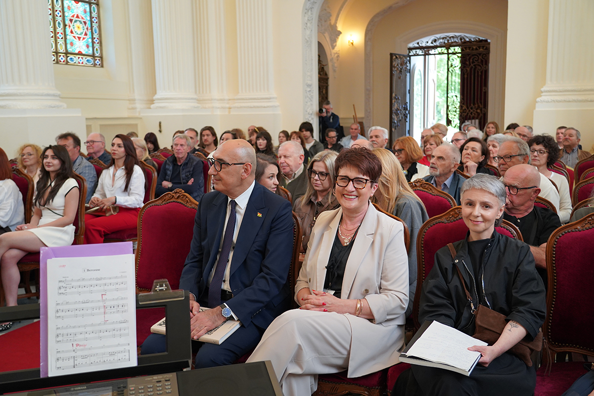 Fotografia przedstawia publiczność zgromadzoną w eleganckiej, zabytkowej sali podczas wydarzenia kulturalnego lub uroczystości. Uczestnicy siedzą na ustawionych w rzędach krzesłach obitych czerwonym, welurowym materiałem, z dekoracyjnymi drewnianymi ramami.
Na pierwszym planie, w dolnej części zdjęcia, widoczny jest fragment instrumentu klawiszowego lub pulpitu muzycznego. Na stojaku znajduje się otwarta partytura z nutami, częściowo zasłaniająca lewą stronę kadru. Sugeruje to, że zdjęcie wykonano z perspektywy osoby występującej lub znajdującej się blisko miejsca koncertowego.
W pierwszym rzędzie siedzą trzy osoby wyraźnie wyróżniające się w kadrze. Po lewej stronie mężczyzna w granatowym garniturze, białej koszuli i ciemnym krawacie patrzy w bok, w kierunku sceny lub prowadzącego wydarzenie. Obok niego siedzi uśmiechnięta kobieta w jasnym, kremowym garniturze, z rękami splecionymi na kolanach. Po prawej stronie znajduje się kobieta z krótkimi, siwymi włosami, ubrana na czarno, trzymająca na kolanach otwarty notes lub zeszyt.
Za nimi widać liczną publiczność w różnym wieku. Część osób patrzy przed siebie, część rozmawia lub spogląda w bok. Wśród uczestników widać zarówno stroje eleganckie, jak i mniej formalne: marynarki, koszule, sukienki, bluzki i okulary przeciwsłoneczne noszone na głowie.
Sala jest jasna i reprezentacyjna. Ściany mają kremowy kolor, a przestrzeń zdobią wysokie kolumny oraz dekoracyjne detale architektoniczne. Po lewej stronie, wysoko na ścianie, znajduje się kolorowy witraż z geometrycznym motywem, w którym wyróżnia się czerwony sześciokąt lub gwiazda. W głębi, po prawej stronie, widoczne są otwarte drzwi z ozdobną, metalową kratą. Przez wejście wpada naturalne światło i zieleń z zewnątrz.
Atmosfera zdjęcia jest uroczysta, ale swobodna. Publiczność wygląda na skupioną i życzliwie nastawioną, a uśmiechy osób w pierwszym rzędzie sugerują pozytywny, kulturalny charakter wydarzenia.