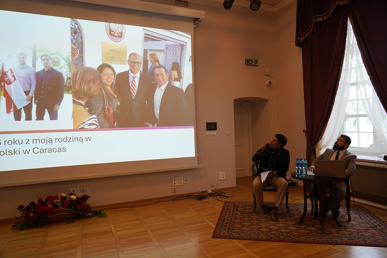 Fotografia przedstawia spotkanie w sali konferencyjnej. Po prawej siedzą dwaj prowadzący; jeden mówi do mikrofonu, drugi obsługuje laptop. Na ekranie wyświetlono zdjęcia związane z Ambasadą Polski w Caracas.