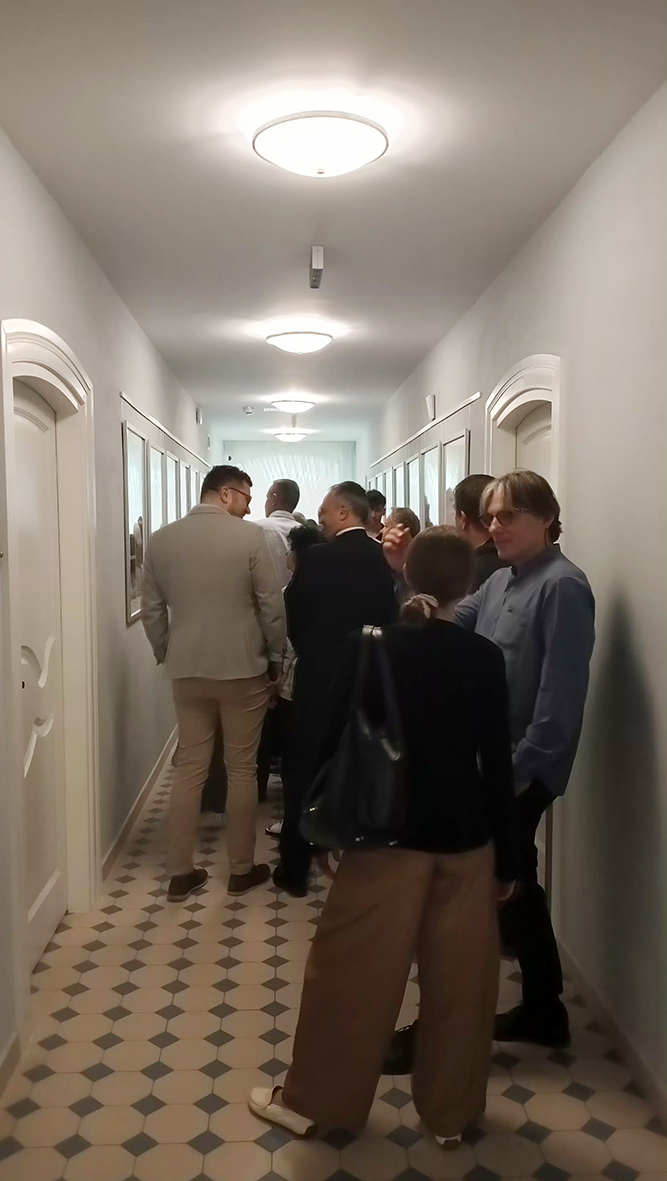 Fotografia przedstawia uczestników wernisażu oglądających wystawę rozmieszczoną w długim, jasnym korytarzu. Ludzie stoją w niewielkich grupach przy pracach zawieszonych na ścianach, rozmawiają i oglądają ekspozycję.