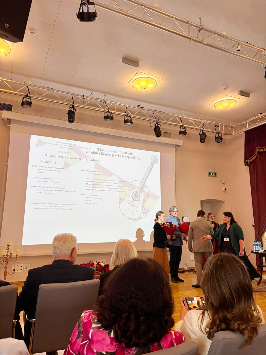 Fotografia przedstawia zakończenie spotkania w sali konferencyjnej. Przed ekranem z programem wydarzenia stoją organizatorzy i uczestnicy, a dwie osoby trzymają bukiety kwiatów. Na pierwszym planie siedzi publiczność,