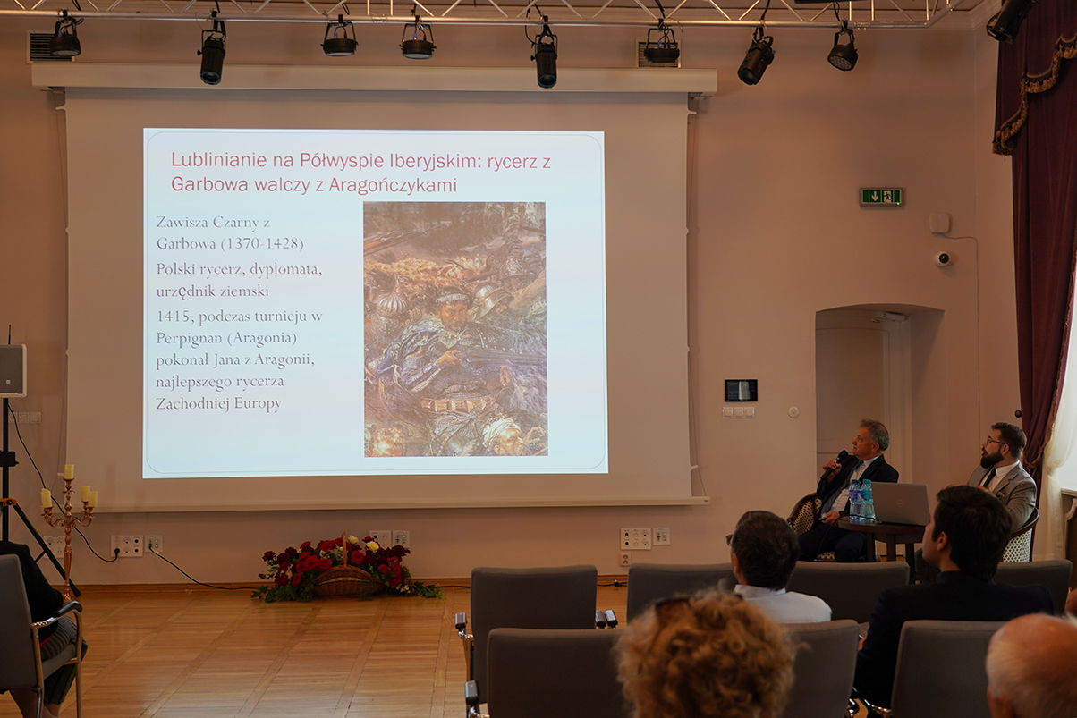 Fotografia przedstawia wykład w sali konferencyjnej. Na ekranie wyświetlono prezentację o Zawiszy Czarnym i jego walkach z Aragończykami. Po prawej stronie siedzą dwaj prowadzący, a przed nimi uczestnicy spotkania.