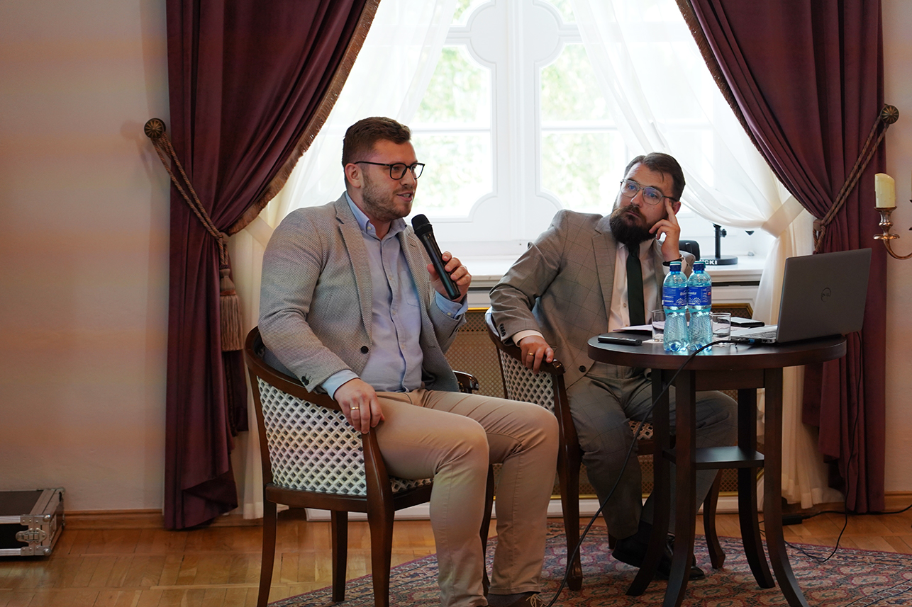 Fotografia przedstawia dwóch mężczyzn prowadzących spotkanie w sali konferencyjnej. Mężczyzna po lewej mówi do mikrofonu, a siedzący obok brodaty mężczyzna go słucha.