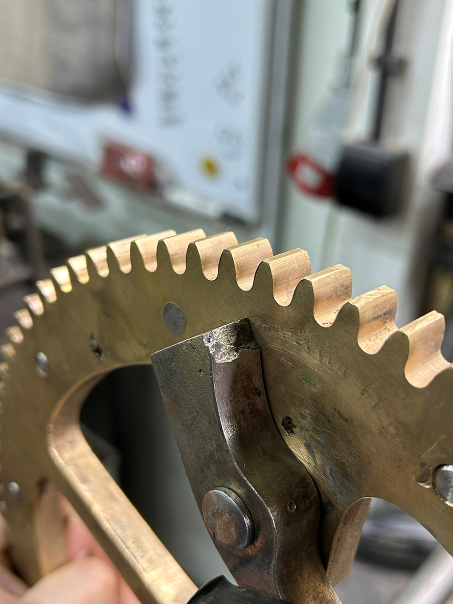 Na fotografii widzimy zbliżenie fragmentu mechanizmu zegara wieżowego. Kadr koncentruje się na dużym, mosiężnym kole zębatym oraz współpracującym z nim ciemniejszym, stalowym elemencie mechanizmu.
Koło ma szerokie, regularnie rozmieszczone zęby. Jego powierzchnia jest matowa, miejscami nosi ślady użytkowania, przebarwienia i drobne zarysowania. Przy krawędzi koła przylega masywny, wyprofilowany stalowy element zakończony niewielką powierzchnią roboczą. W jego dolnej części widoczny jest okrągły nit lub sworzeń.
Po lewej stronie kadru znajduje się kolejne mosiężne koło mechanizmu, częściowo przesłonięte i lekko nieostre. W dolnym lewym rogu widoczna jest dłoń podtrzymująca element konstrukcji.
Tło pozostaje mocno rozmyte. Można dostrzec w nim jasne ściany pracowni oraz fragmenty wyposażenia technicznego. Dzięki niewielkiej głębi ostrości uwaga skupia się przede wszystkim na precyzyjnie wykonanych zębach koła oraz styku współpracujących części mechanizmu.
Fotografia ukazuje detal zabytkowego mechanizmu zegarowego w trakcie prac konserwatorskich i podkreśla precyzję jego konstrukcji.