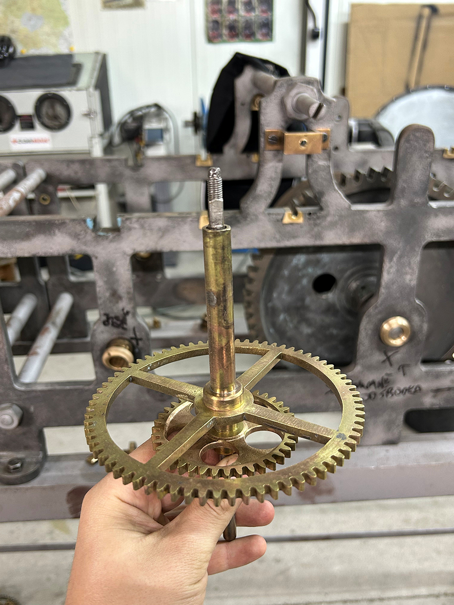 Na fotografii widzimy zbliżenie elementu mechanizmu zegara wieżowego trzymanego w dłoni. Na pierwszym planie znajduje się zespół kilku mosiężnych kół zębatych osadzonych na pionowym wale.

Największe koło ma formę szerokiego pierścienia z drobnymi, równomiernie rozmieszczonymi zębami na obwodzie. Jego środek łączą z osią cztery wąskie ramiona. Poniżej widoczne jest mniejsze koło zębate z kilkoma okrągłymi otworami. Cały zespół ma złocistobrązowy kolor charakterystyczny dla mosiądzu. Na powierzchni metalu widoczne są ślady użytkowania i delikatne przebarwienia.
Przez środek mechanizmu przebiega długi pionowy wał, zakończony u góry gwintowanym trzpieniem. Dłoń podtrzymuje element od dołu, dzięki czemu dobrze widoczna jest jego konstrukcja i sposób połączenia poszczególnych części.
W tle znajduje się znacznie większa, stalowa rama mechanizmu zegara wieżowego z otworami, osiami i fragmentami kolejnych kół zębatych. Tło jest lekko rozmyte, dlatego uwaga skupia się na trzymanym w dłoni mosiężnym zespole.
Fotografia dokumentuje jeden z etapów prac konserwatorskich i pozwala z bliska zobaczyć precyzję wykonania elementów dużego mechanizmu zegarowego.